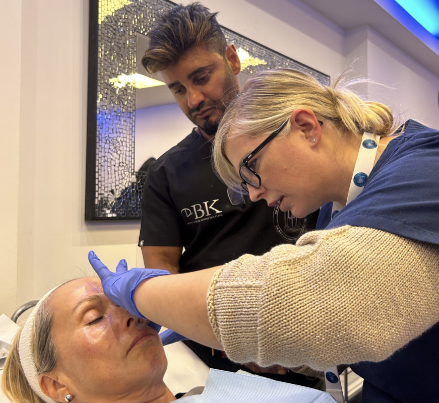 A female medical professional wearing glasses administering a cosmetic treatment to a woman lying down. A male practitioner in black scrubs observing closely. 